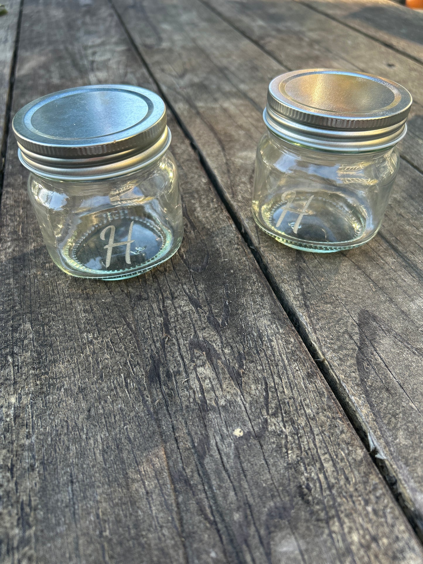 Two glass jars with metal lids on a wooden surface engraved with initial
